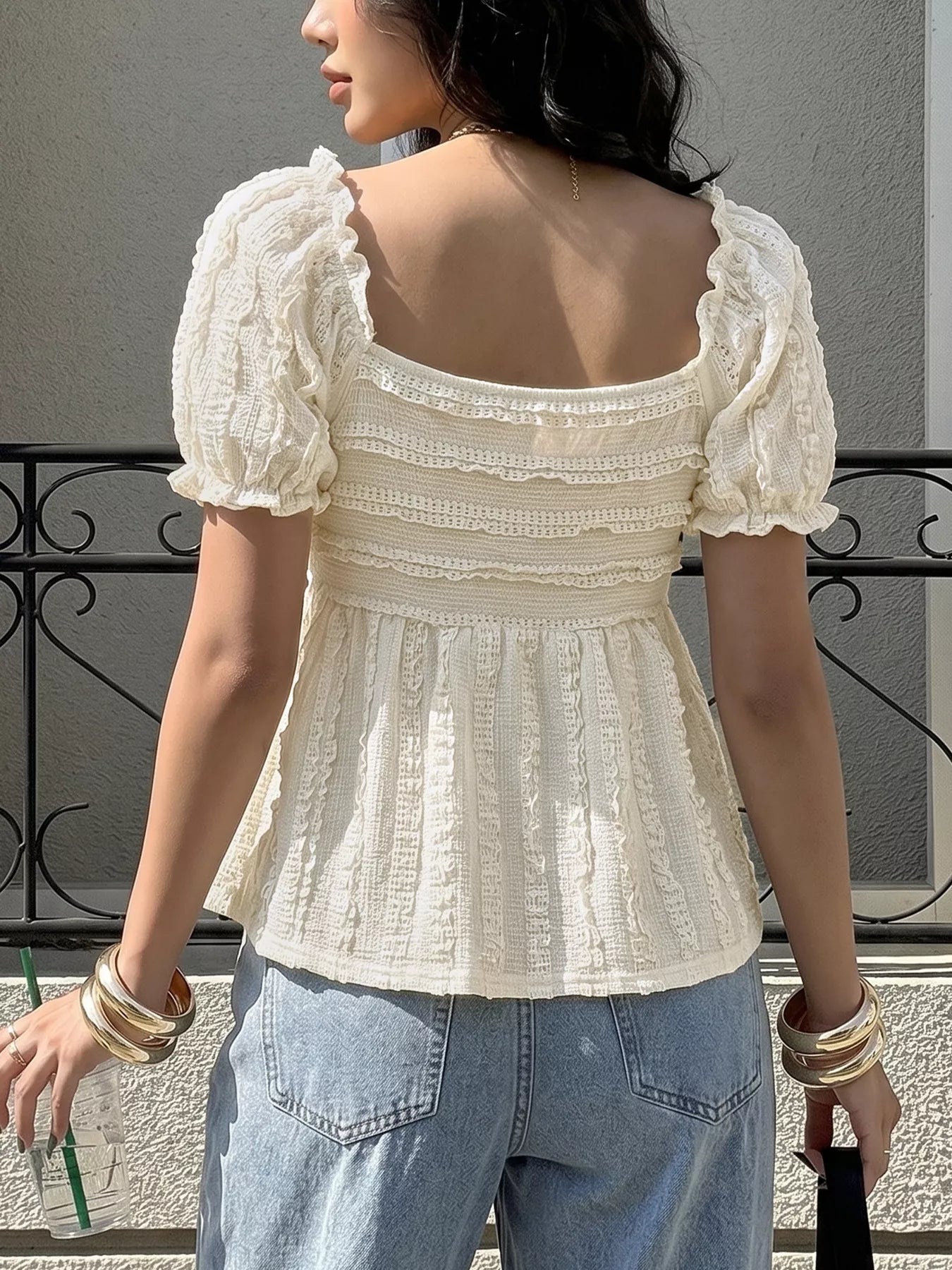 Woman wearing a white lace top with puffed sleeves and blue jeans, standing against a neutral background.