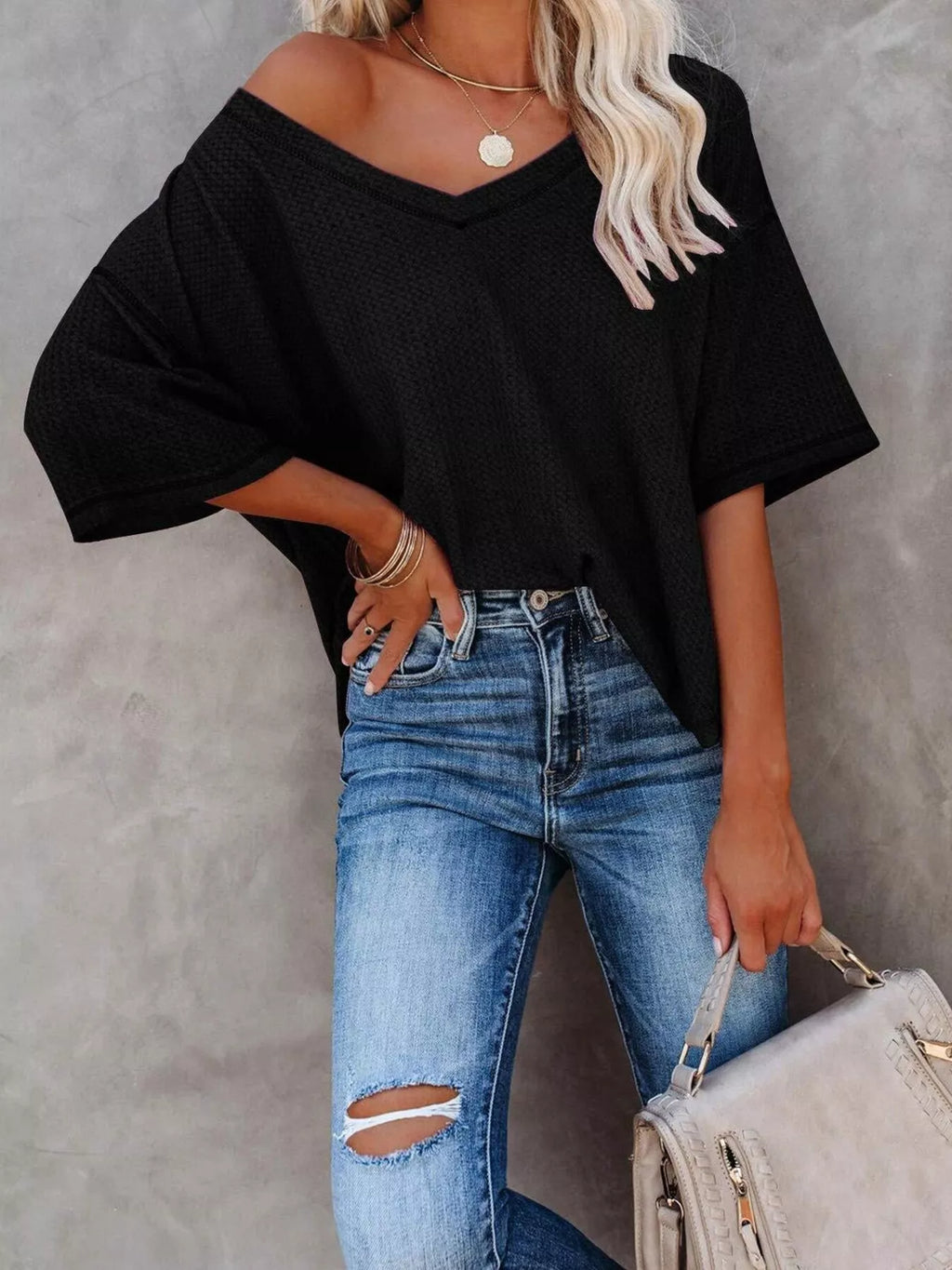 Person wearing a black off-shoulder top and blue jeans, holding a beige handbag against a gray background.