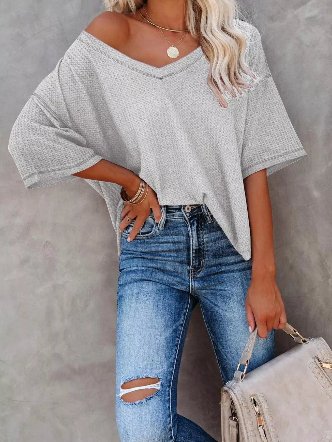 Woman wearing a gray off-shoulder top and blue jeans, holding a beige handbag against a gray background.