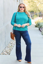 Woman wearing a teal top and blue jeans standing on a sidewalk.