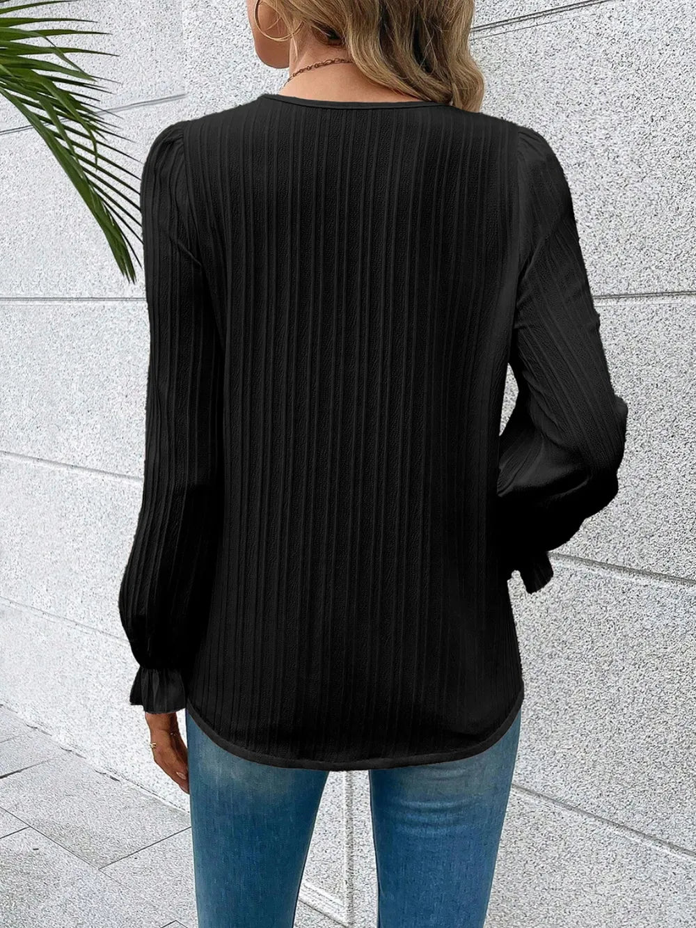 Person wearing a black pleated blouse with long sleeves against a light gray wall.
