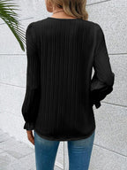 Person wearing a black pleated blouse with long sleeves against a light gray wall.
