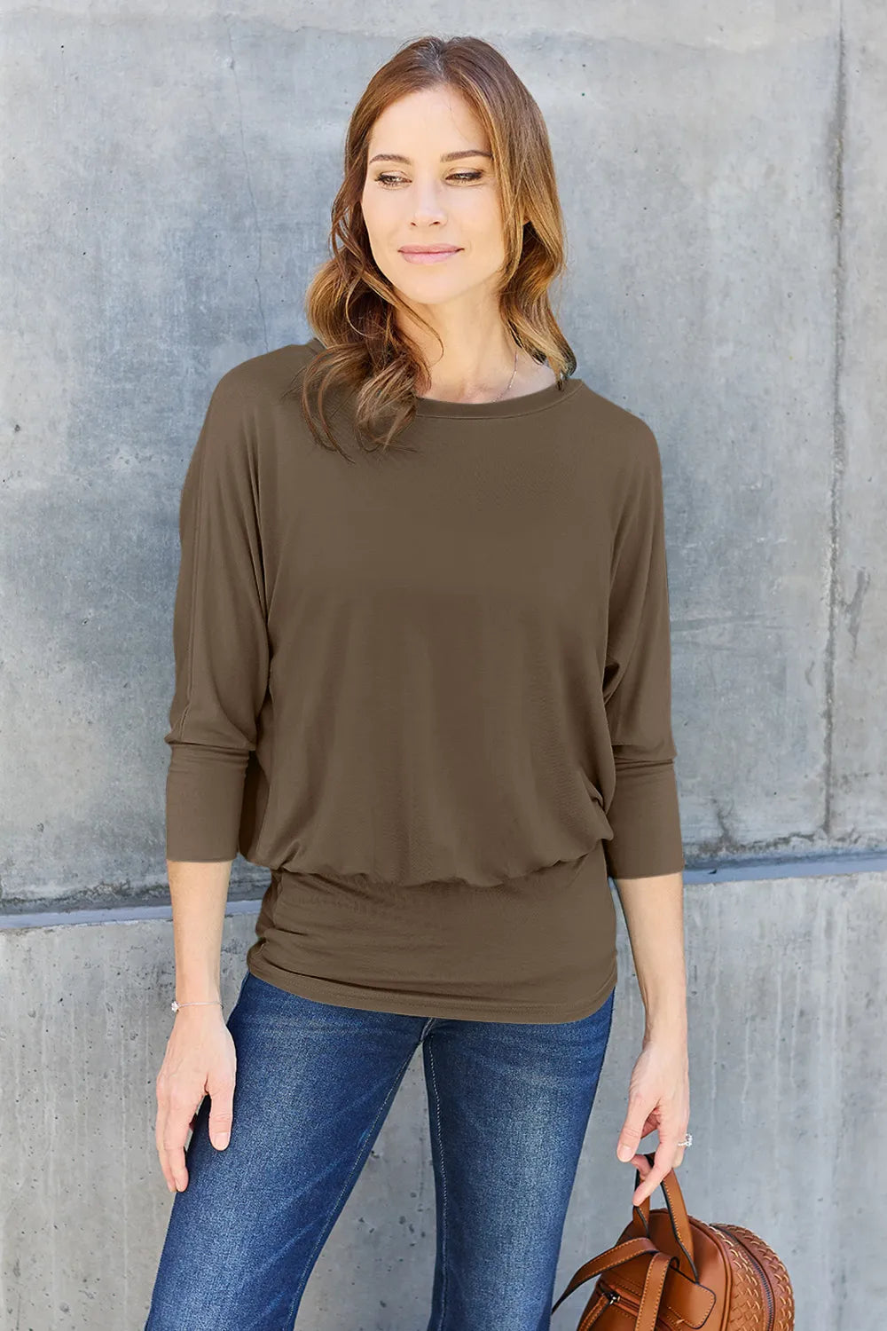 Woman wearing an olive green top and blue jeans against a gray concrete wall.