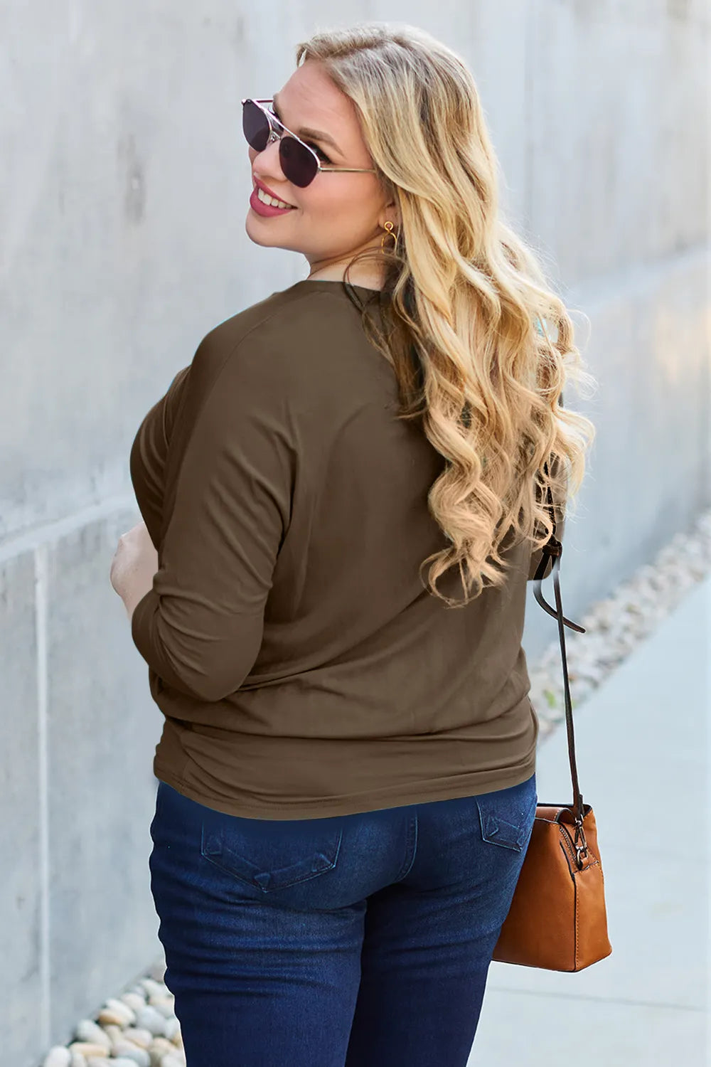 Woman wearing a brown long-sleeve top and blue jeans, holding a brown handbag against a light-colored wall.