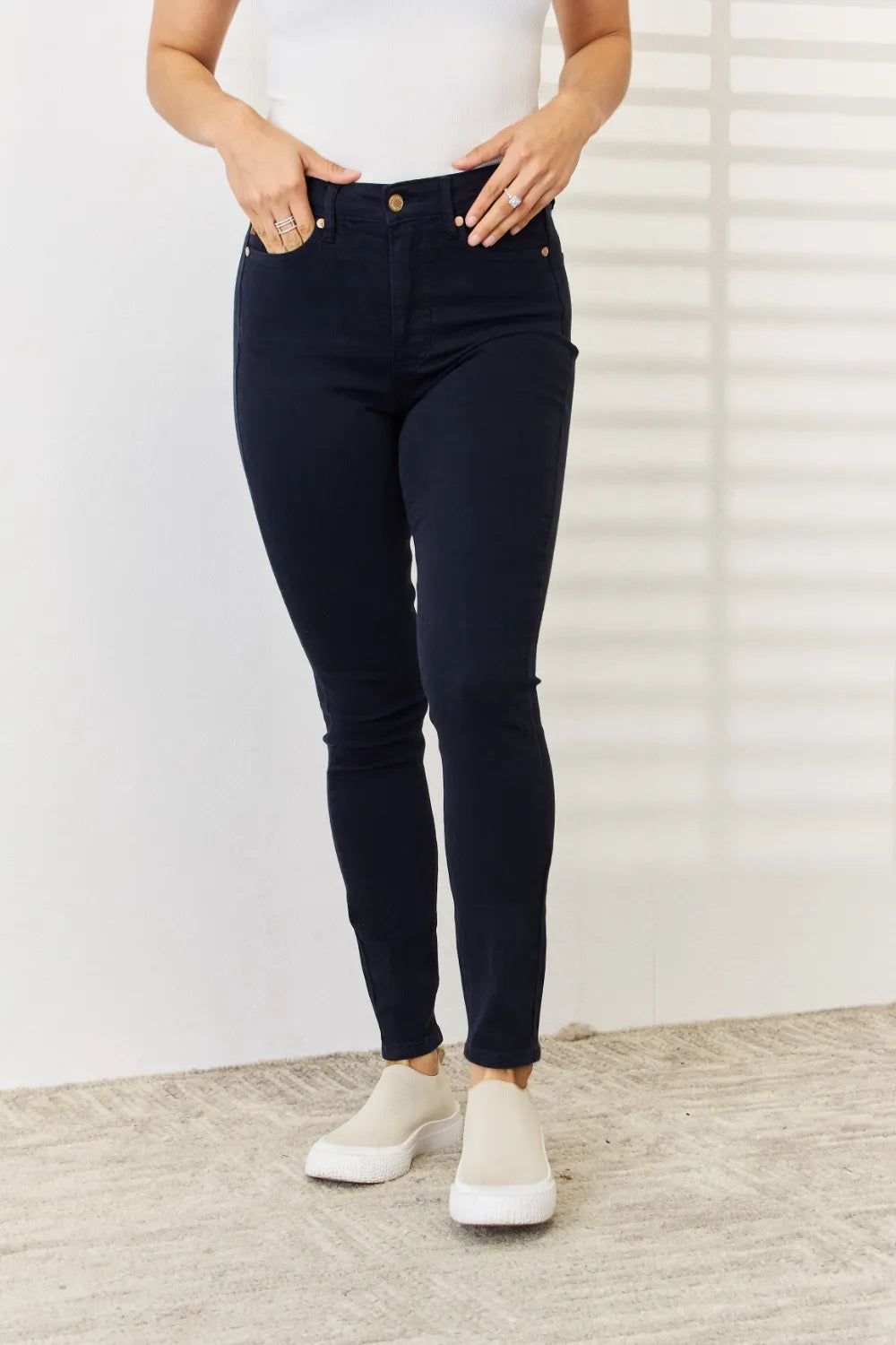Person wearing dark blue jeans and white sneakers against a white wall.