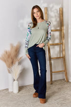 Woman wearing a green top with floral sleeves and blue jeans standing in a room with a wooden ladder and decorative plants.