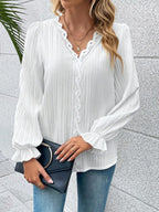 Woman wearing a white blouse with long sleeves and blue jeans, holding a black clutch against a light gray wall.