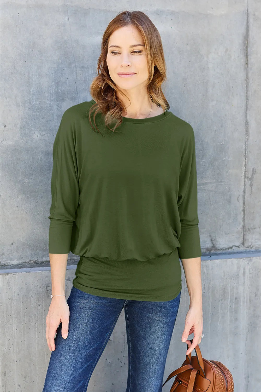 Woman wearing an olive green top and blue jeans against a gray wall.