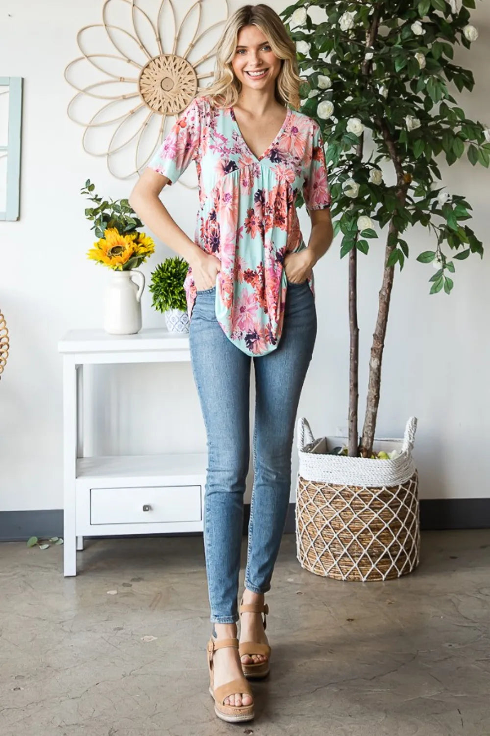 Woman wearing a floral blouse and jeans standing in a room with plants and decor.
