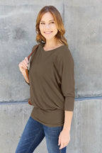 Woman wearing a brown long-sleeve top and blue jeans against a gray concrete wall.