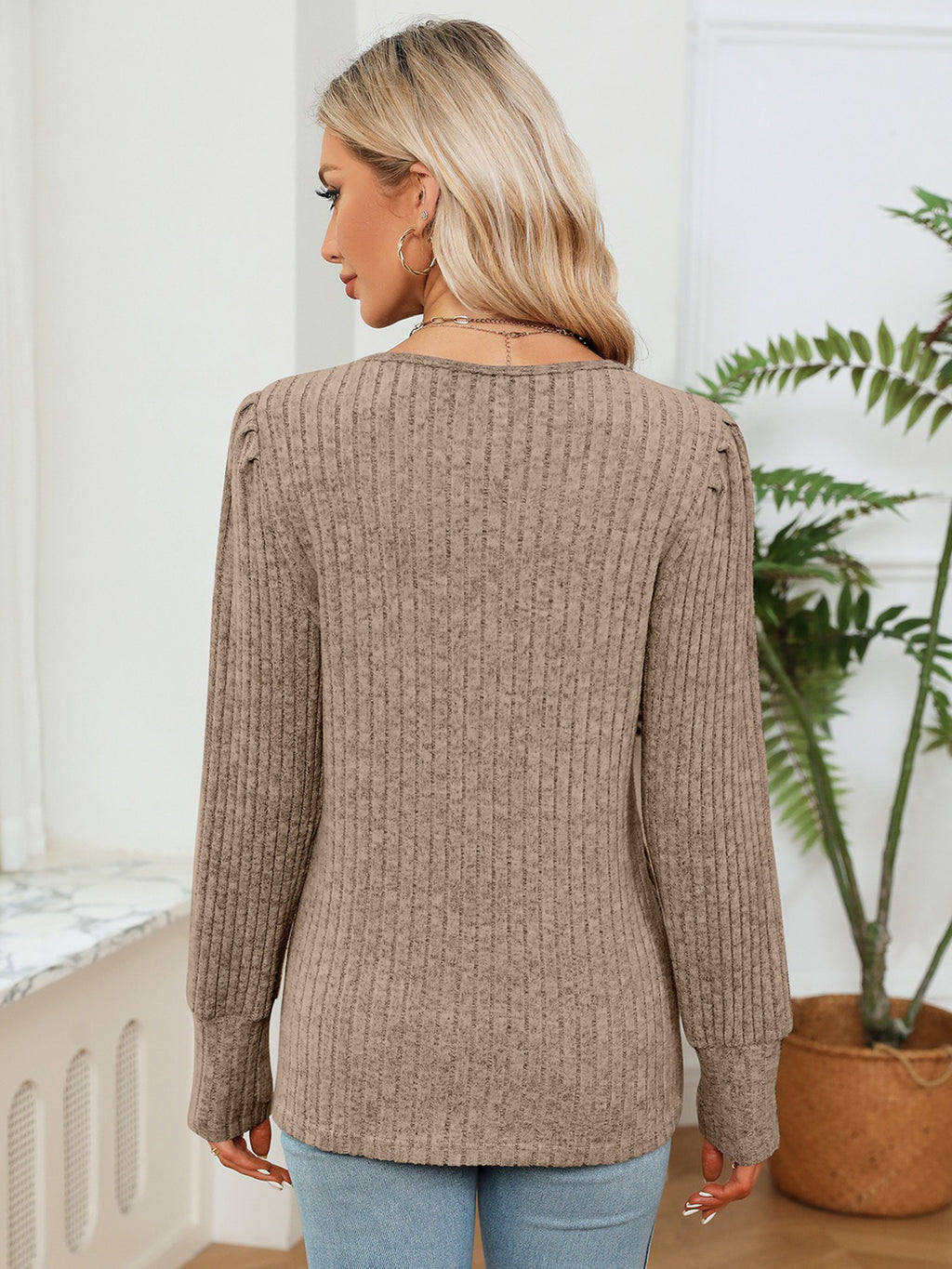 Woman wearing a brown ribbed sweater in a room with a plant and window.