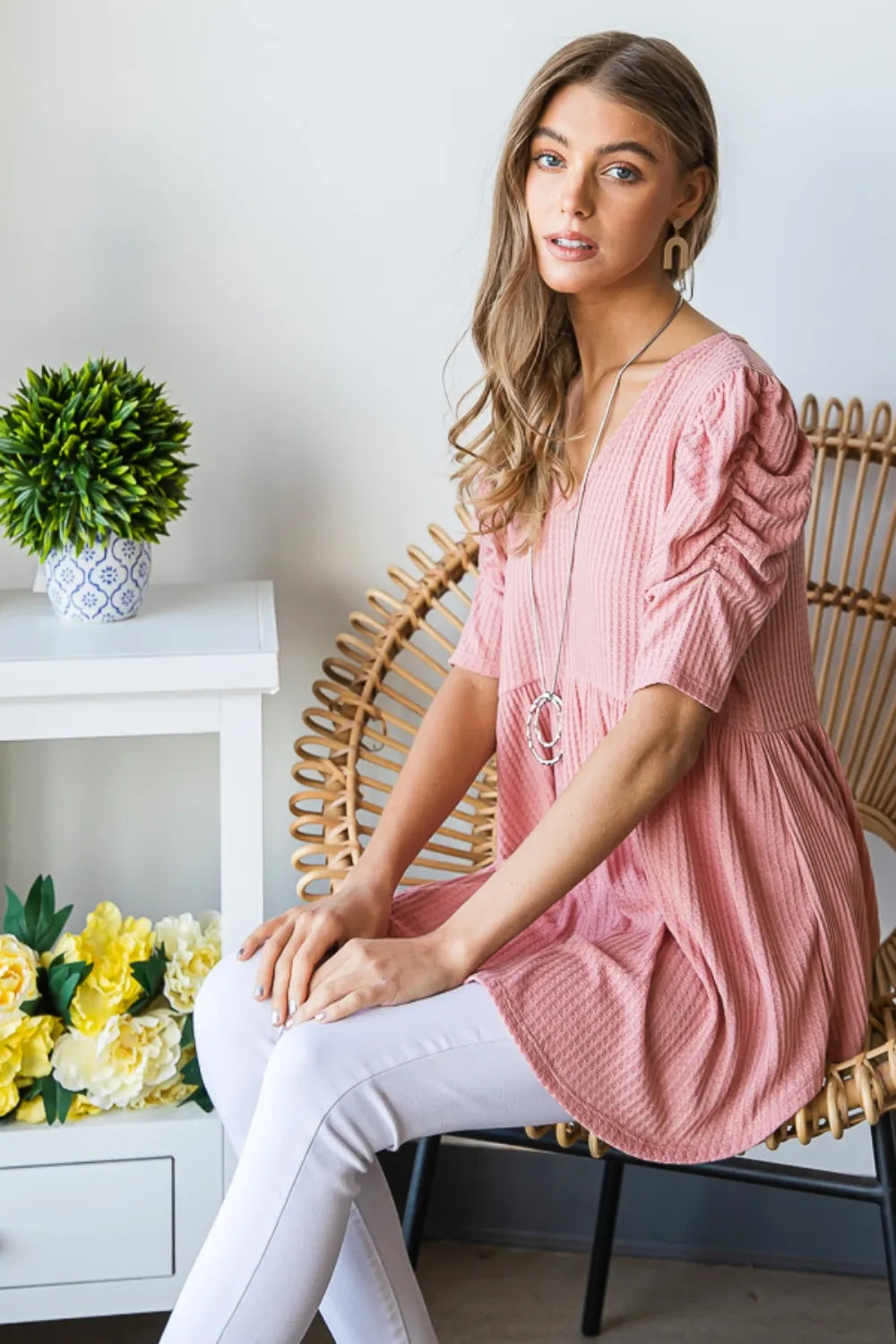 Woman in a pink dress sitting on a wicker chair with a white wall and decorative items in the background