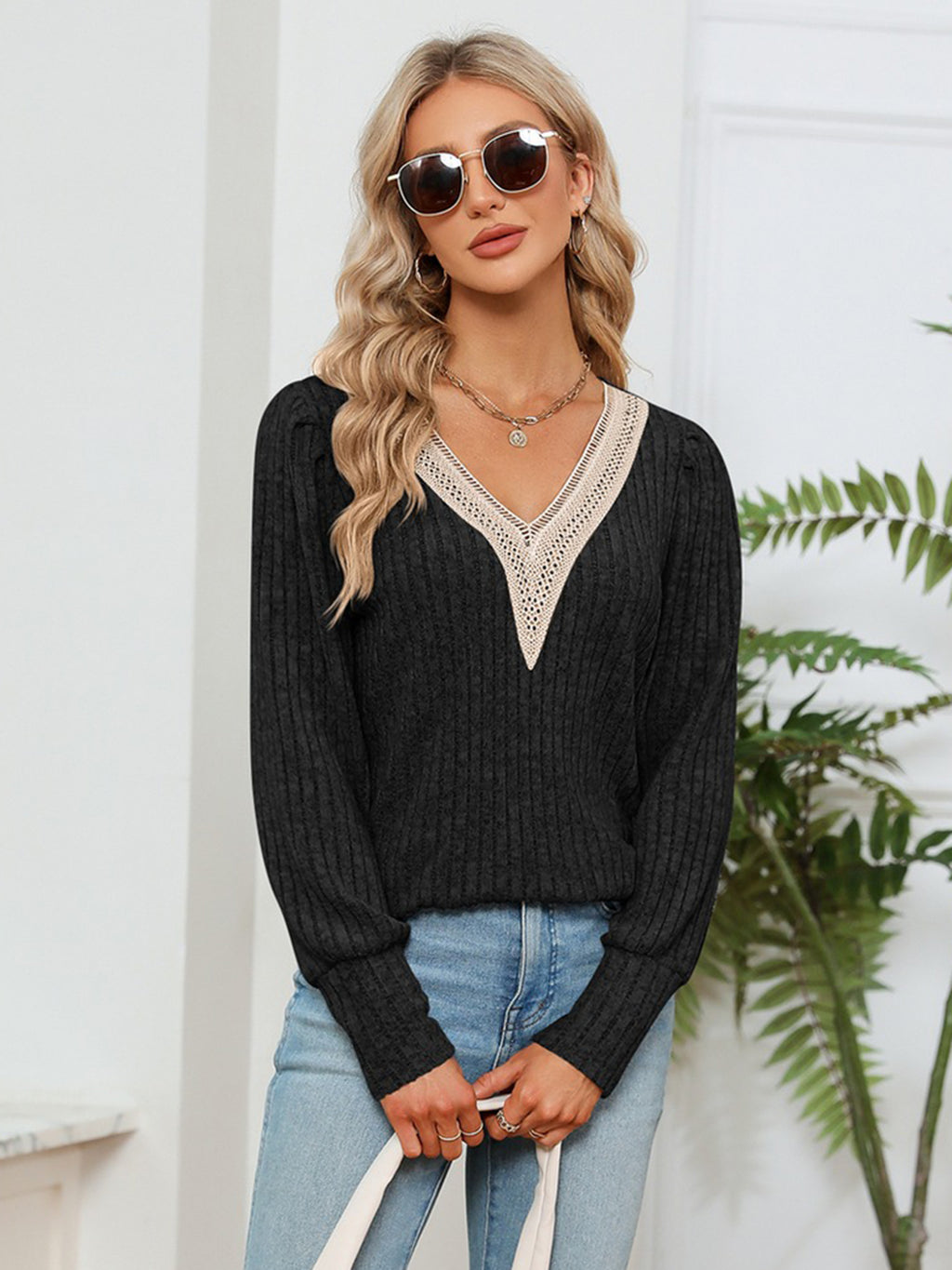 Woman wearing a black sweater with lace details and blue jeans, standing indoors with a plant in the background.