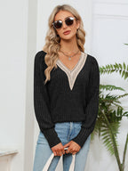 Woman wearing a black sweater with lace details and blue jeans, standing indoors with a plant in the background.