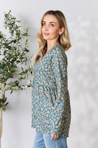 Woman wearing a blue floral long-sleeve top against a white background with a plant to the left.