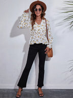 Woman wearing a floral blouse, black pants, and a brown hat against a white wall.