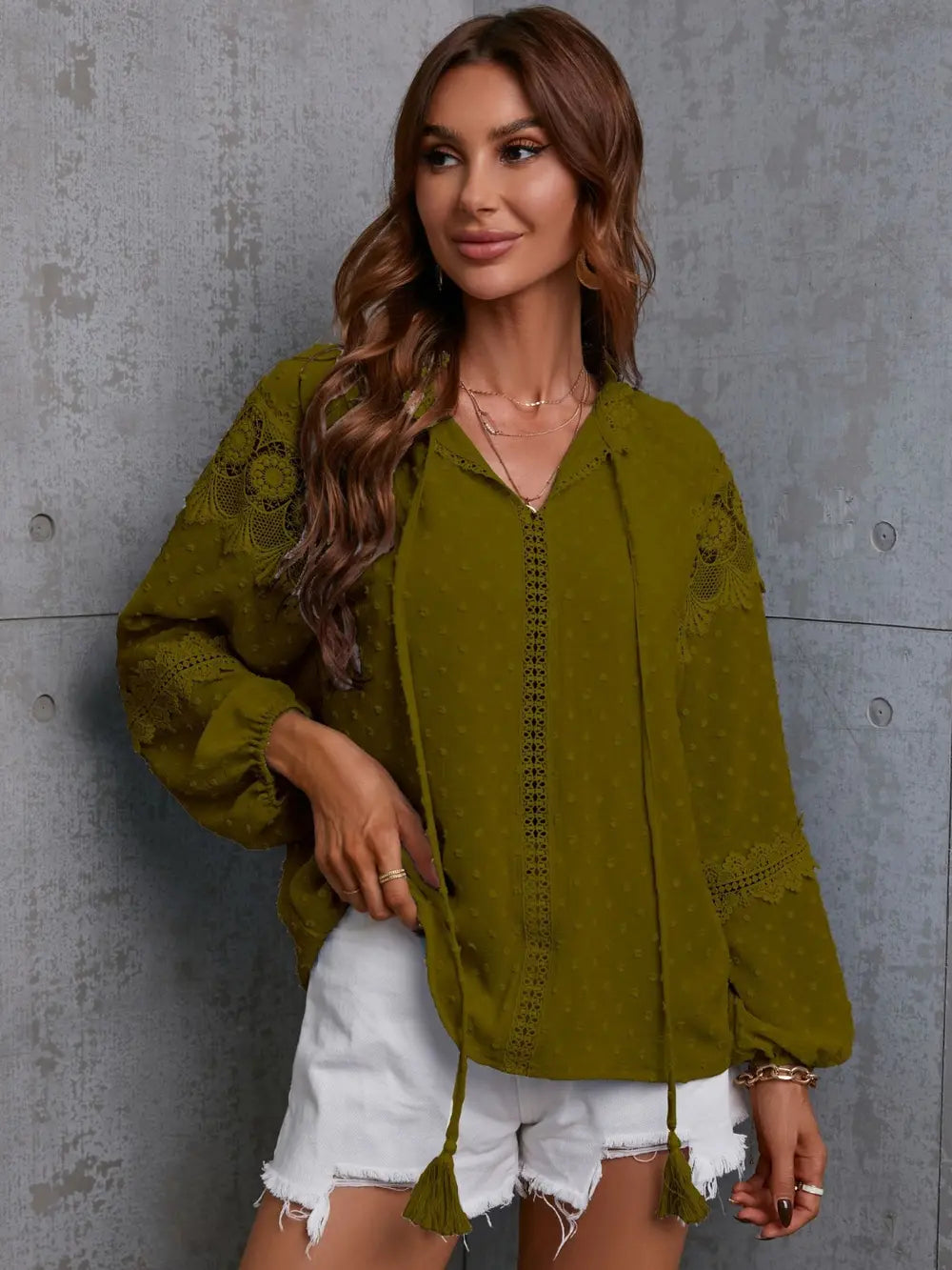 Woman wearing a green embroidered blouse and white shorts against a gray concrete wall.