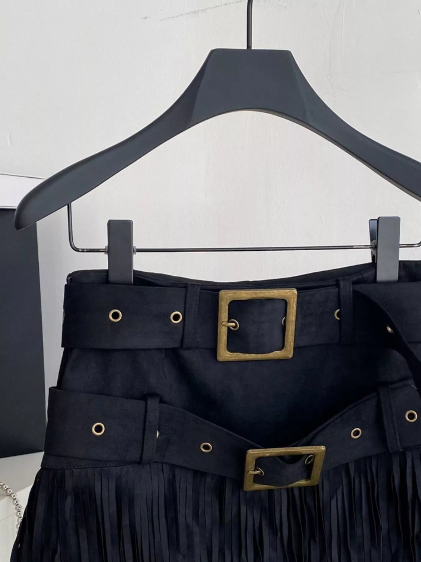 Black fringe skirt with gold buckles on a hanger against a white background