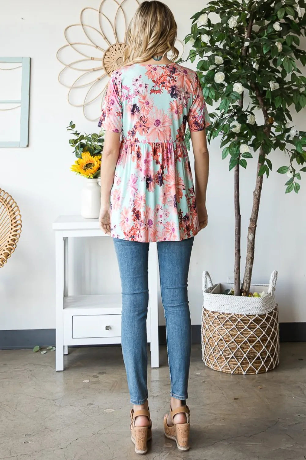 Person wearing a floral blouse and blue jeans standing in a room with a plant and table.