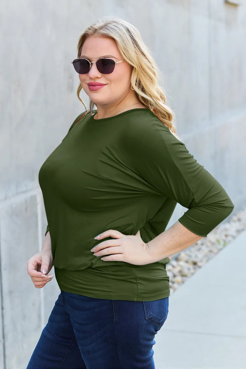 Woman wearing an olive green top and sunglasses against a light gray wall.