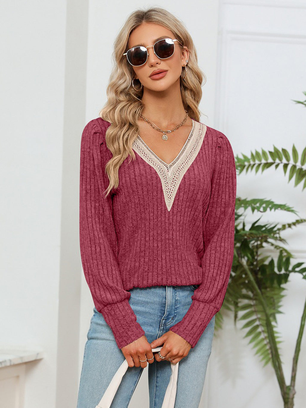Woman wearing a maroon sweater with lace trim and light blue jeans, standing against a white wall with a plant.