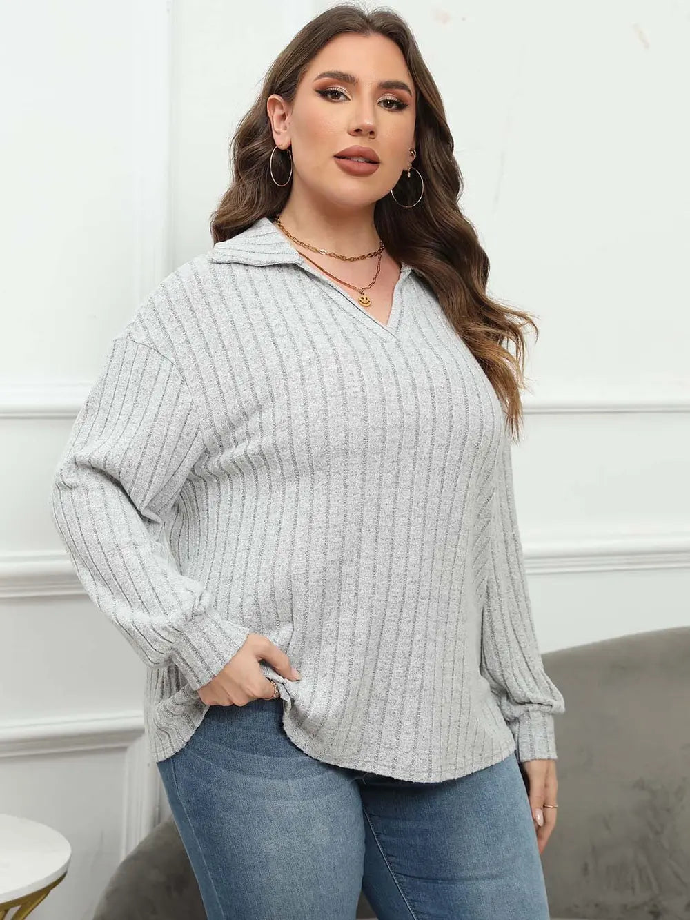 Woman wearing a light gray ribbed sweater and blue jeans against a white wall.